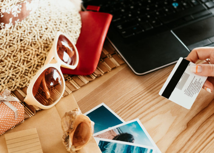 Table filled with travel gear, sunglasses, a laptop, and a woman's hand holding a credit card